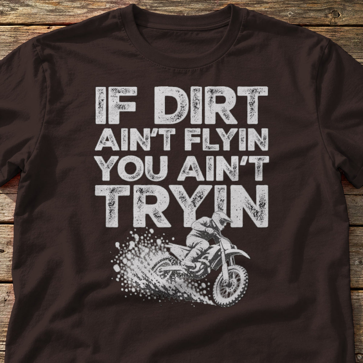 If Dirt Ain't Flyin You Ain't Tryin t-shirt in Dark Chocolate, laid flat on weathered wood
