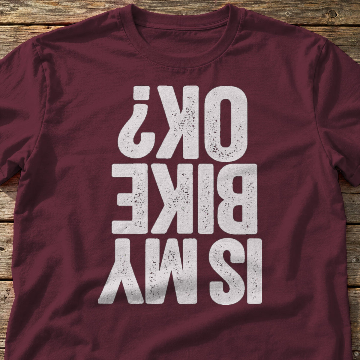 Is My Bike OK? t-shirt in Maroon, laid flat on weathered wood