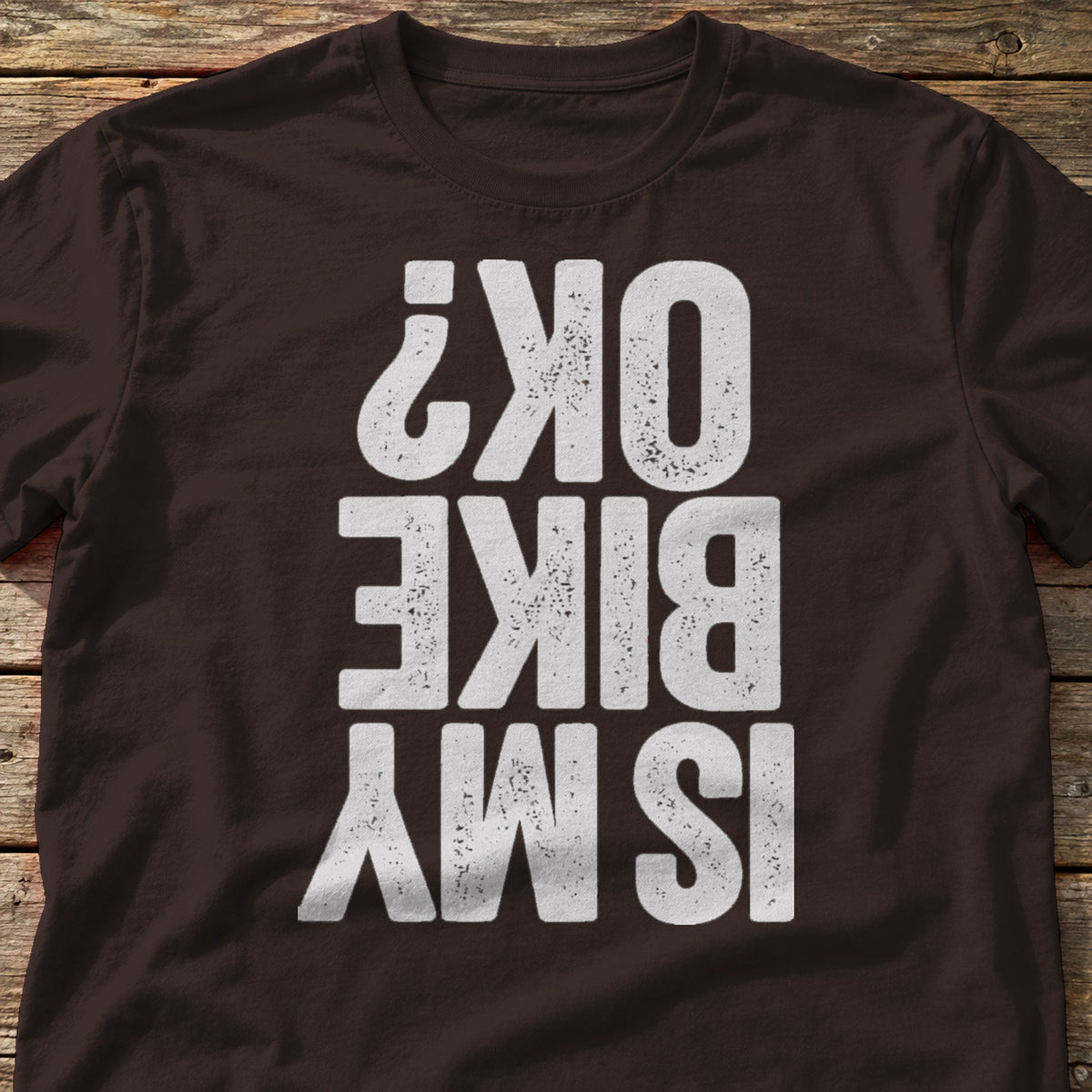 Is My Bike OK? t-shirt in Dark Chocolate, laid flat on weathered wood